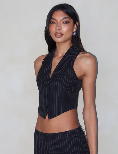 A person with long dark hair is dressed in a fashionable TAMARA VEST - BLACK : PINSTRIPE from TIGERMIST, paired with matching pants, standing against a light gray background. They have a confident and poised expression, with one hand resting by their side.