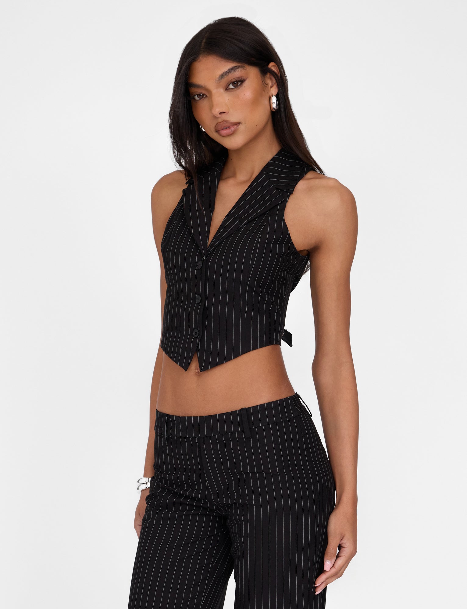 A woman with long, dark hair is standing against a plain white background, wearing the TIGERMIST TAMARA VEST - BLACK : PINSTRIPE along with matching tailored-fit pinstripe pants. She is looking directly at the camera with her hands by her sides and is accessorized with earrings and bracelets.