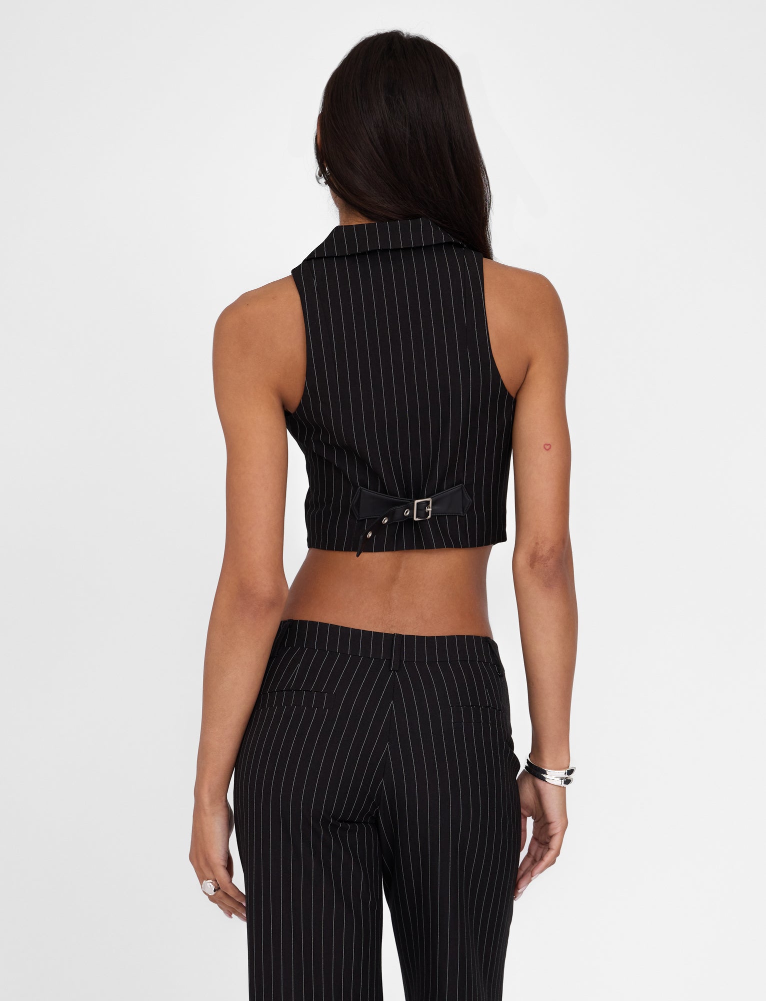 A person with long dark hair is seen from behind wearing the TAMARA VEST - BLACK : PINSTRIPE by TIGERMIST and matching high-waisted pants, both showcasing a tailored fit. The buckle detail at the back of the vest adds a touch of sophistication against the plain white background.