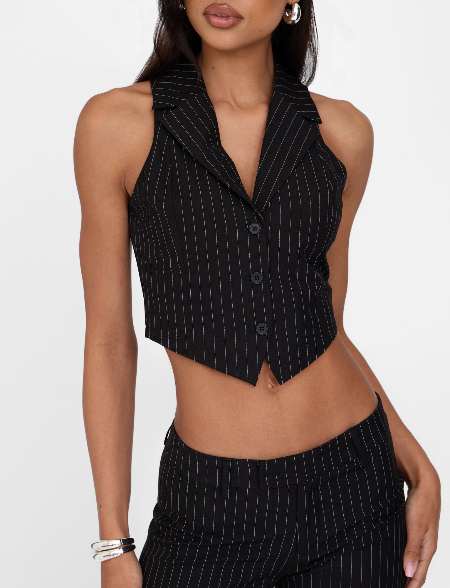 A person dressed in the TIGERMIST TAMARA VEST - BLACK : PINSTRIPE, showcasing its stylish sleeveless design, deep V-neckline, and button-up front. They have paired this tailored-fit vest with matching black pinstriped trousers and accessorized with bracelets and hoop earrings.