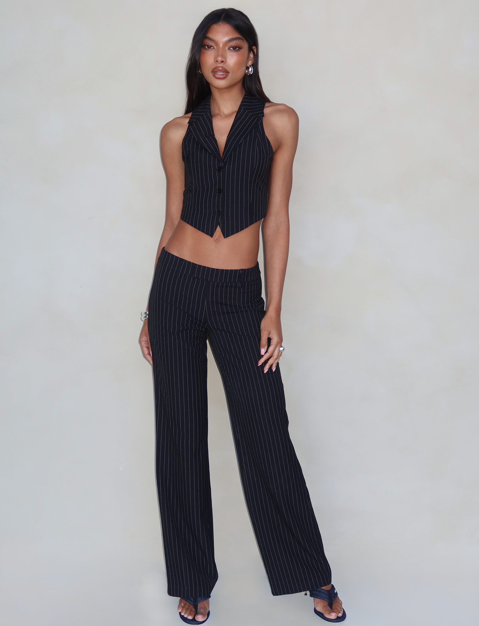A person with long dark hair stands against a plain background, donning a black pinstripe vest paired with the TIGERMIST TAMARA PANT - BLACK : PINSTRIPE, which features a high-waisted, wide-leg design. They complete the look with dark open-toe heels and minimal jewelry, including earrings and bracelets.