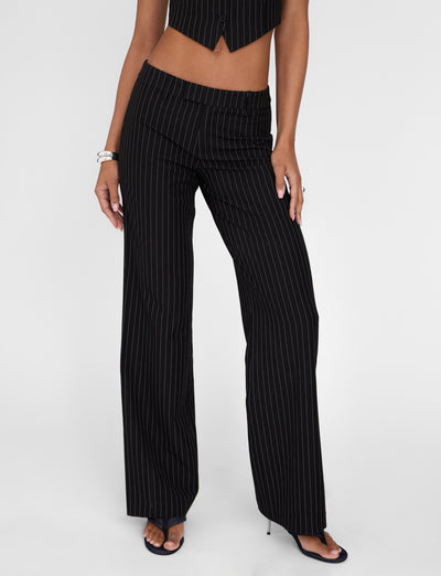 A person is seen wearing the TIGERMIST TAMARA PANT in black pinstripe with wide-leg design and a mid-rise waist, paired with a matching cropped vest. The outfit is completed with black open-toe heels and accessorized with a silver bracelet. The image captures only the lower half of the body against a plain white background.