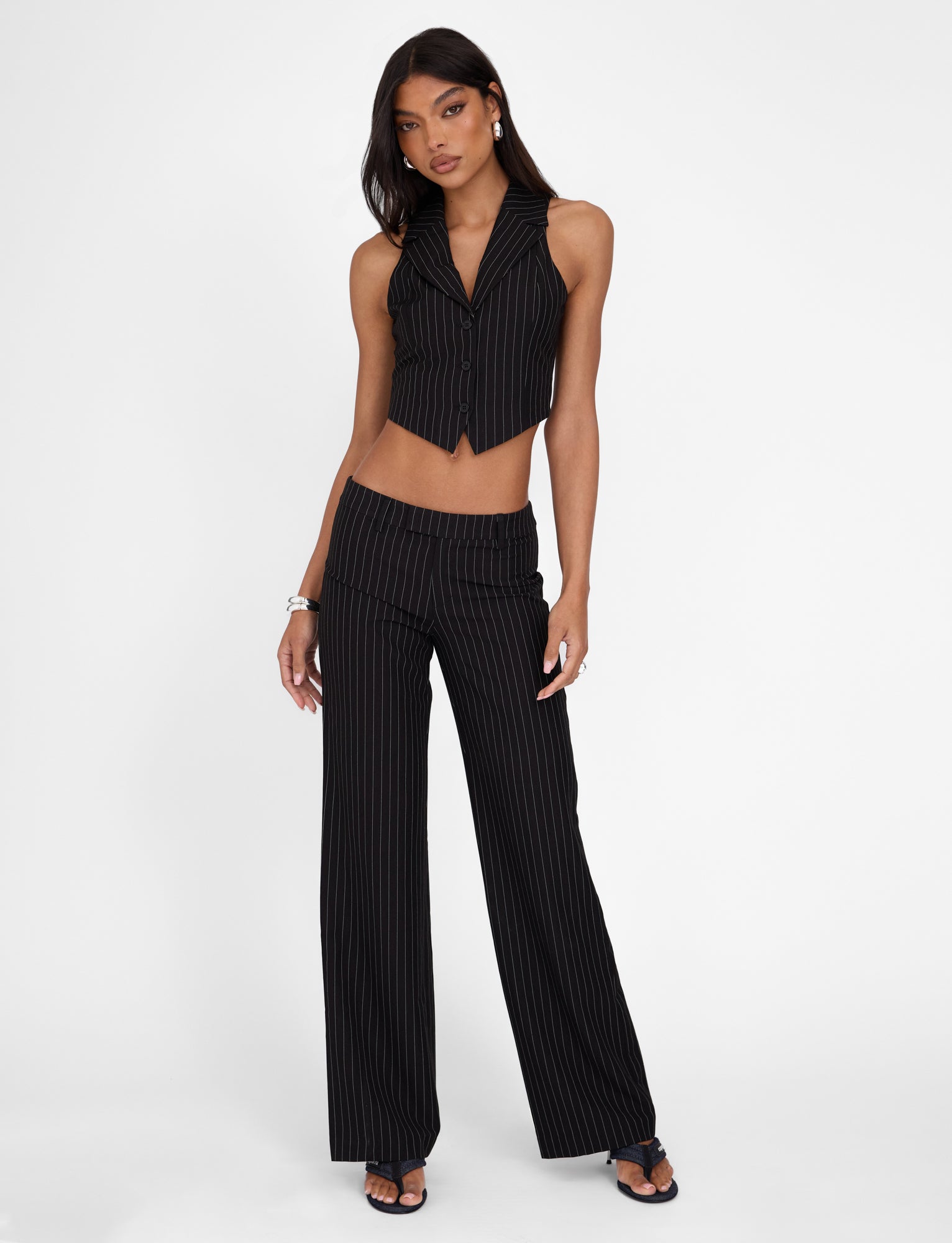 A woman wearing the TIGERMIST TAMARA PANT - BLACK: PINSTRIPE and a matching vest stands against a white background. She has long dark hair and accessorizes with a bracelet, earrings, and black sandals. The mid-rise waist outfit from Tigermist reveals her midriff, and she poses with one hand on her hip.