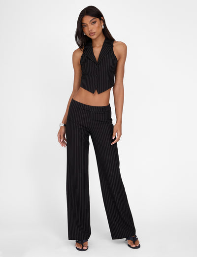 A woman wearing the TIGERMIST TAMARA PANT - BLACK: PINSTRIPE and a matching vest stands against a white background. She has long dark hair and accessorizes with a bracelet, earrings, and black sandals. The mid-rise waist outfit from Tigermist reveals her midriff, and she poses with one hand on her hip.