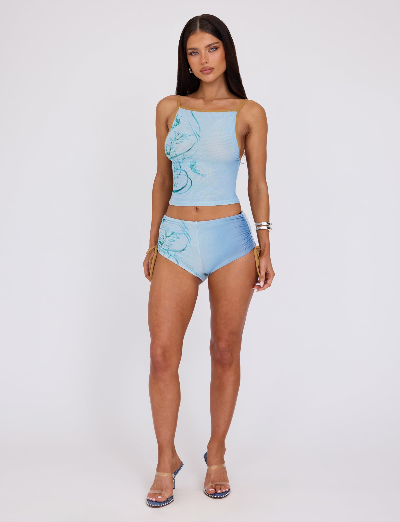 A woman stands confidently against a plain background, donning the ELVERA SHORT - BLUE : OMBRE by TIGERMIST. Her outfit includes a light blue, sleeveless top and matching high-waisted shorts adorned with a subtle floral pattern. She has long, straight hair and accessorizes with bracelets and clear high-heeled sandals.