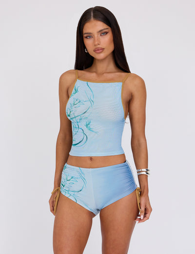A woman with long, dark hair stands against a plain background, wearing the fitted silhouette of the TIGERMIST ELVERA TOP - BLUE : GRADIENT FLORAL crop top paired with high-waisted shorts. The outfit features a subtle floral design, and she accessorizes with silver hoop earrings and a stack of silver bracelets on her right wrist.