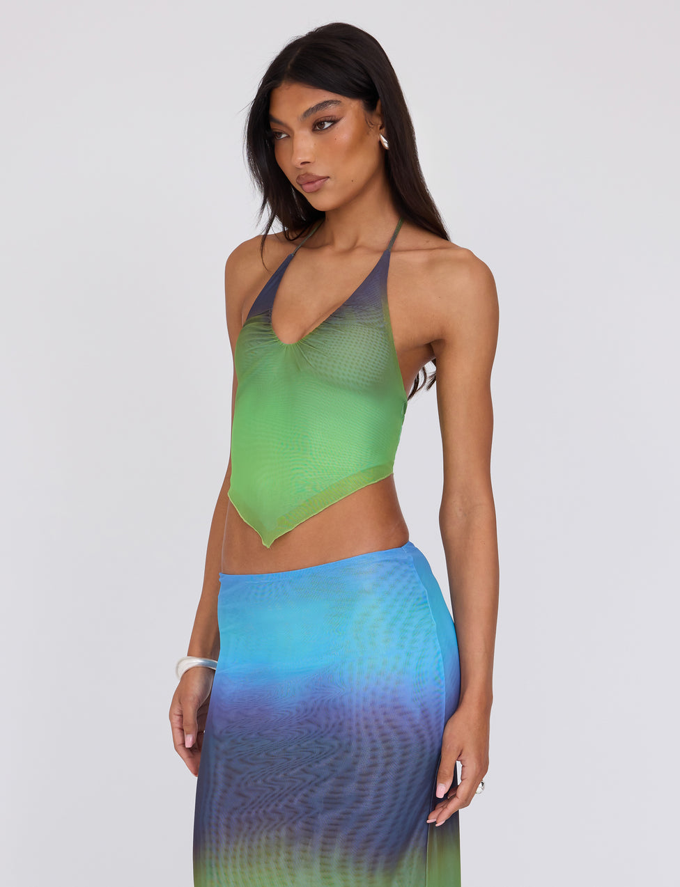 A woman with long, dark hair is wearing the NOELY TOP - MULTI : OMBRE : GRADIENT STRIPE from TIGERMIST, along with a matching skirt. She has a serious expression and stands facing slightly to the left against a plain white background. Her jewelry is minimal, including just a bracelet.