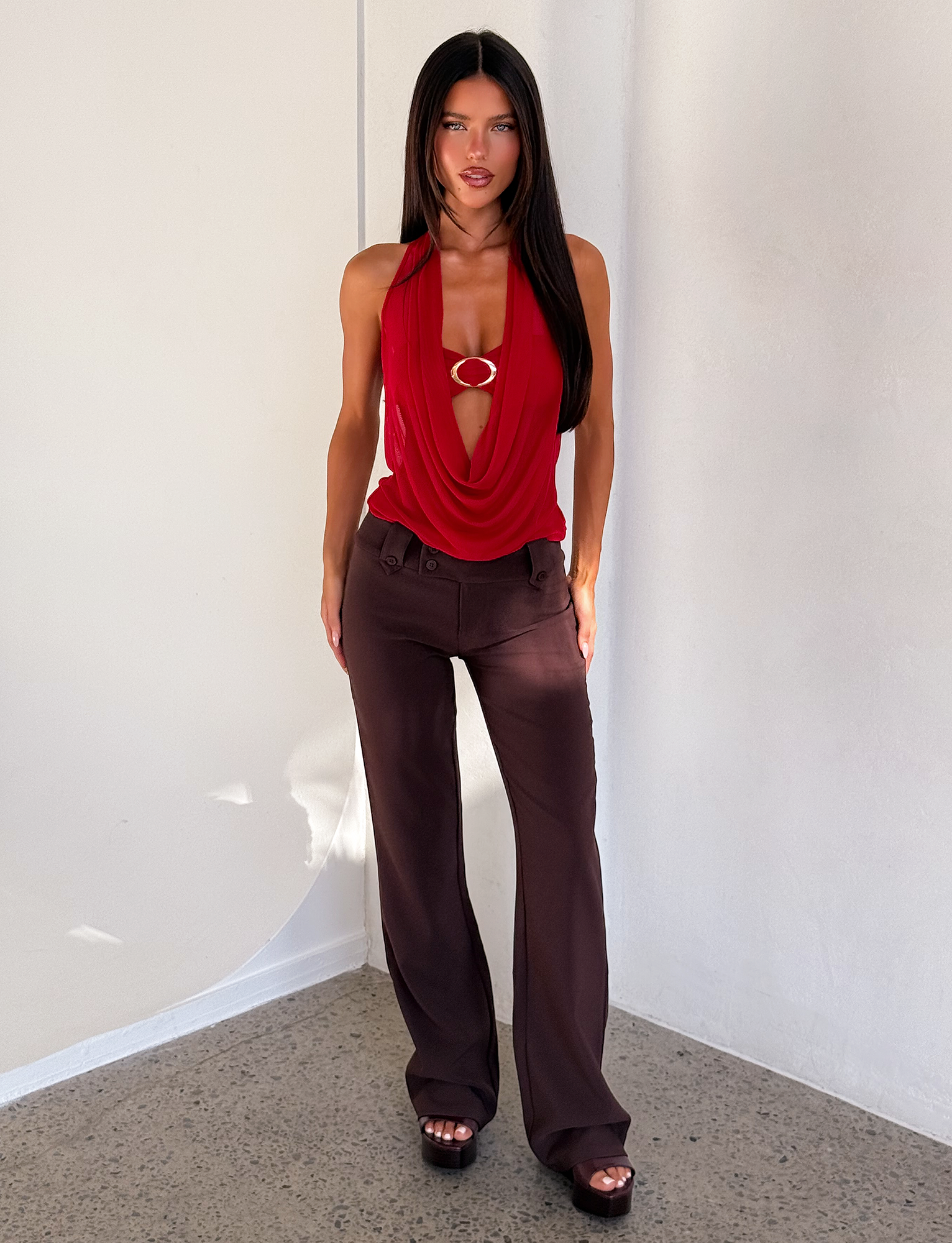 A woman with long dark hair wears the TIGERMIST MAKENA TWO-PIECE TOP in red and high-waisted dark brown pants, standing indoors against a white wall on a speckled floor.