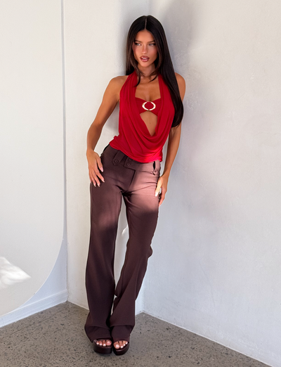 A woman with long dark hair stands against a white wall, wearing the TIGERMIST MAKENA TWO-PIECE TOP in red with a circular gold detail, brown wide-leg pants, and dark platform sandals. She looks straight at the camera.