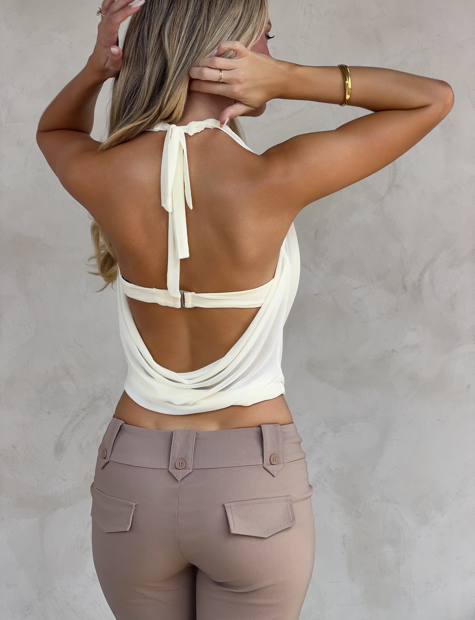 A woman with long blonde hair stands against a light textured wall, wearing the TIGERMIST MAKENA TWO-PIECE TOP in ivory and high-waisted beige pants, her back to the camera and one hand touching her head.