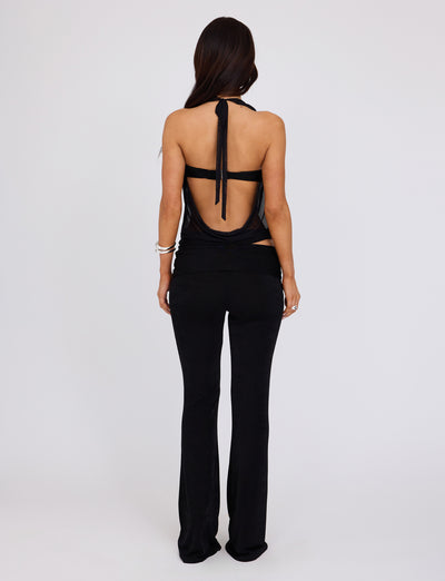 A person with long dark hair is standing with their back to the camera against a plain white background. They are wearing a black, backless halter top and TIGERMIST's MAKENA PANT in black. The top is tied at the neck with a strap, and they have a bracelet on their left wrist.