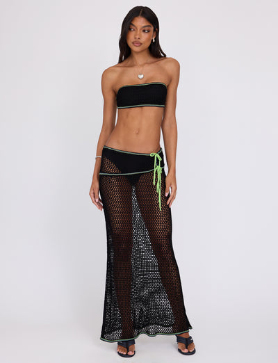 A woman stands modeling Tiger Mist's AZARIA TOP in black, pairing it with a matching black bikini bottom and a long black mesh skirt adorned with neon green accents. She completes her look with black sandals and a silver necklace featuring a heart pendant, posing against a plain white background.
