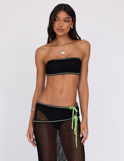 A woman is posing against a plain white background, wearing the Tiger Mist AZARIA TOP - BLACK, a matching black crochet bandeau top with green trim, paired with a skirt cover-up. She has long, dark hair and accessorizes with a heart necklace and earrings. Her expression is neutral as she looks slightly to the side.
