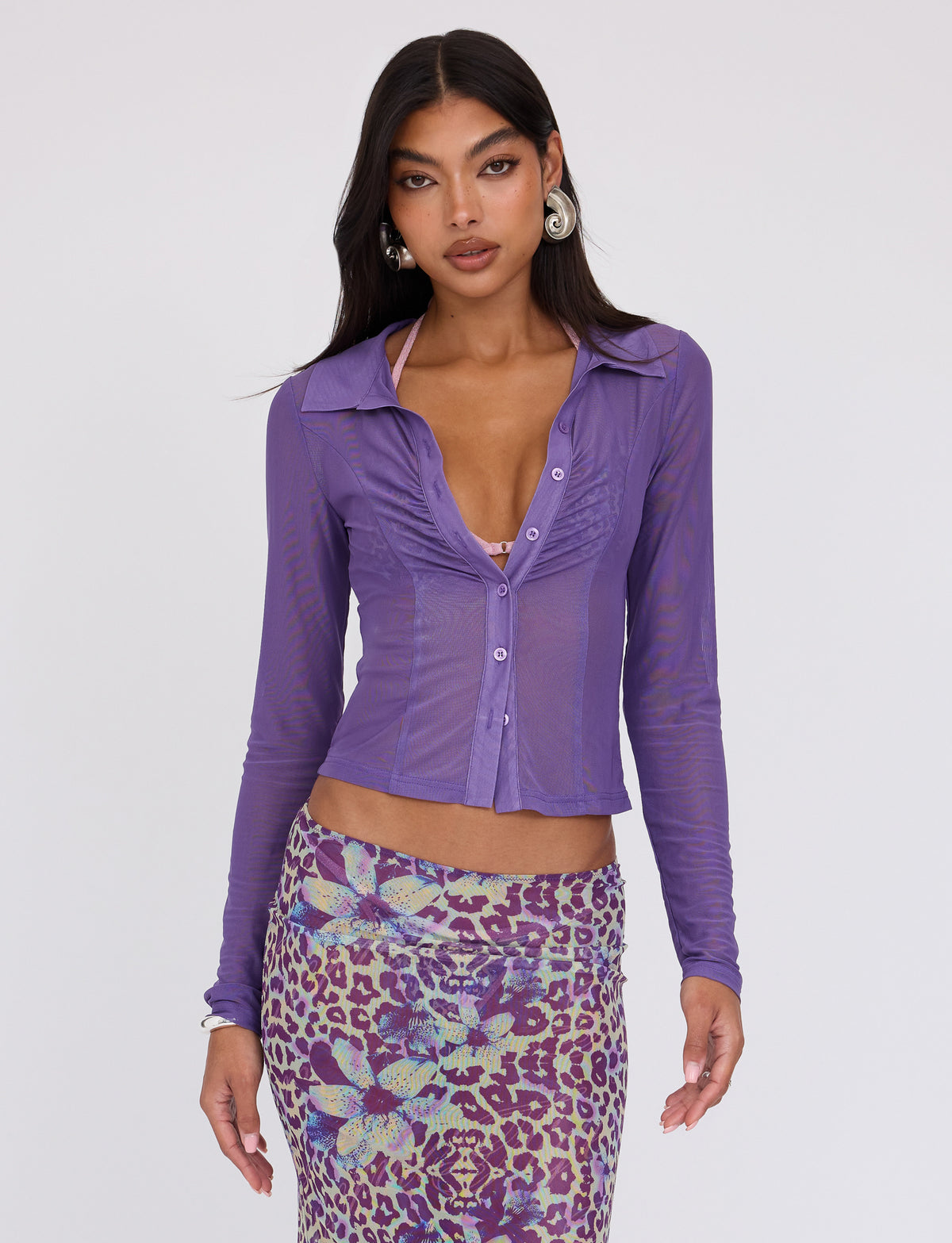 A woman with long dark hair dons the AHLING LONG SLEEVE TOP in purple from TIGERMIST, paired with a colorful, patterned skirt that features a mix of leopard and floral designs. She poses against a plain white background with a confident expression.
