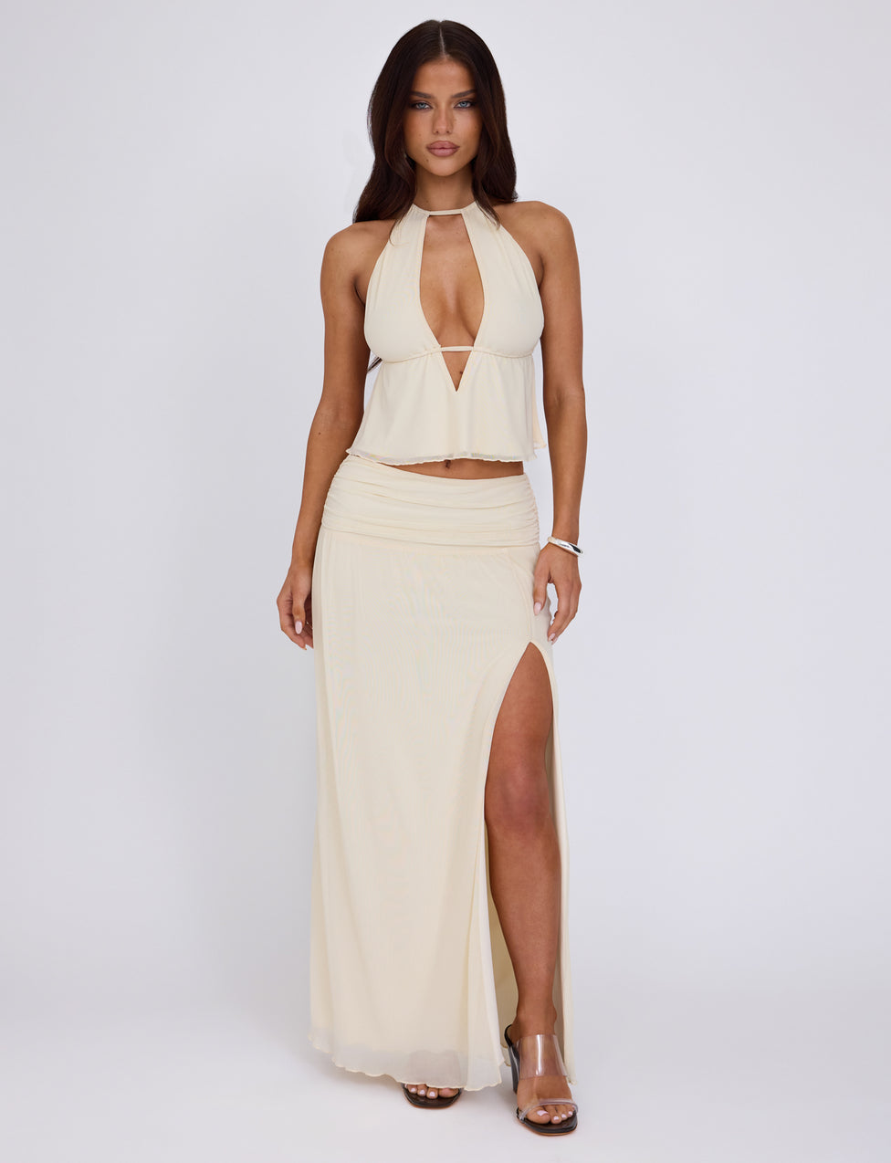 A woman stands against a plain white background, wearing the BIRDEE SKIRT in White and Cream from TIGERMIST. The skirt features an asymmetrical high slit that reveals one leg, paired with a light beige halter-top dress boasting a deep neckline. She accessorizes with a bracelet and clear high-heeled sandals, her hair down and flowing loosely.