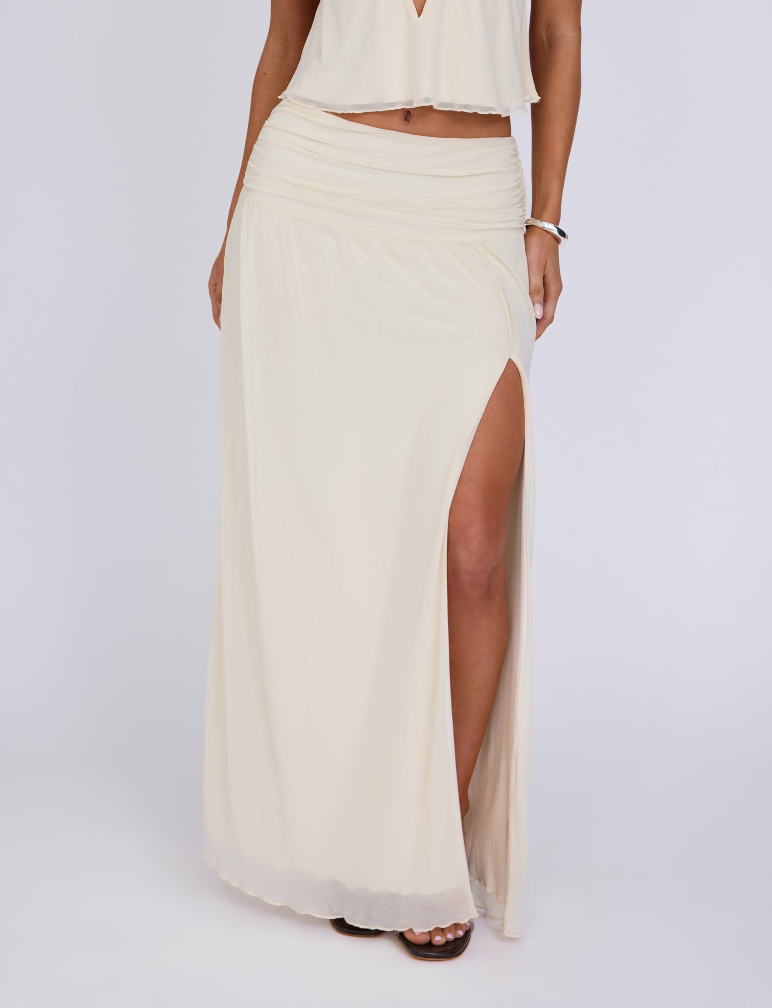 A person wearing a flowing, light cream Tiger Mist Birdee Skirt with a thigh-high slit on the side, standing against a plain white background. The skirt is paired with a matching crop top, and a bracelet is visible on the person's left wrist.
