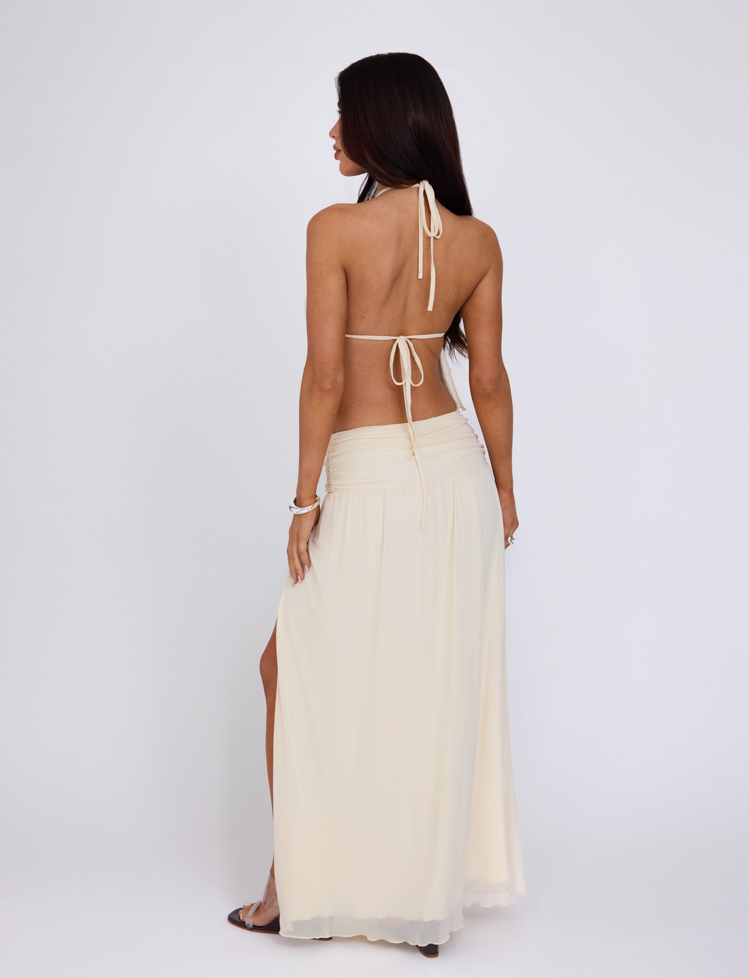 A woman with long, dark hair is seen from behind, wearing a cream-colored, backless halter top and the BIRDEE SKIRT - WHITE : CREAM from TIGERMIST. The flowing skirt features a slit and she stands against a plain white background with her hands relaxed at her sides. She also wears a silver bracelet on her left wrist.