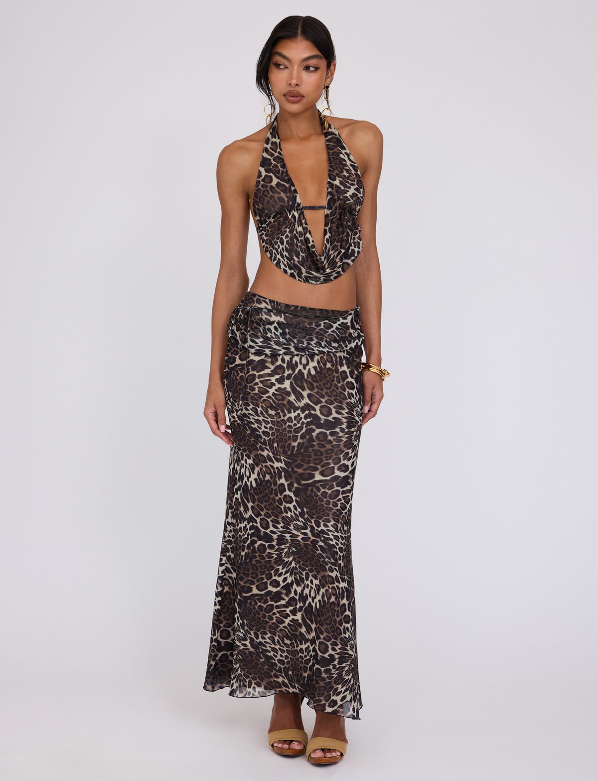 A woman poses against a plain background wearing the TIGERMIST PEARL MAXI SKIRT - MULTI : LEOPARD with a matching halter-neck top, styled with gold earrings, a bracelet, and brown high-heeled sandals.