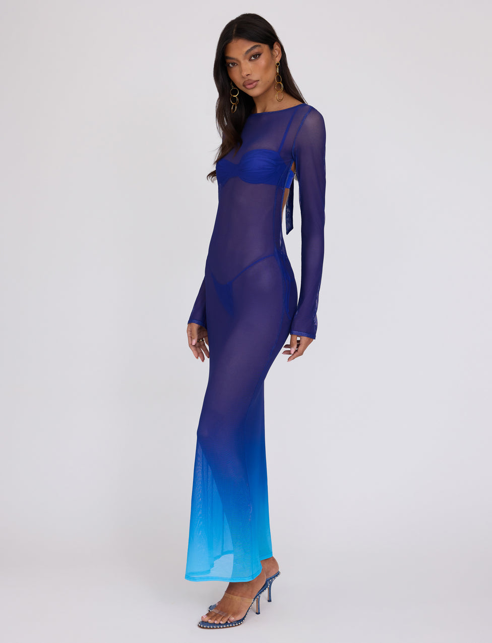 A woman stands against a plain background, wearing the MEILANI DRESS - BLUE: OMBRE from TIGERMIST. The dress is sheer and fades into a lighter blue at the bottom. She has long, dark hair and is posed slightly sideways, looking at the camera. She completes the look with blue high-heeled sandals and gold earrings.