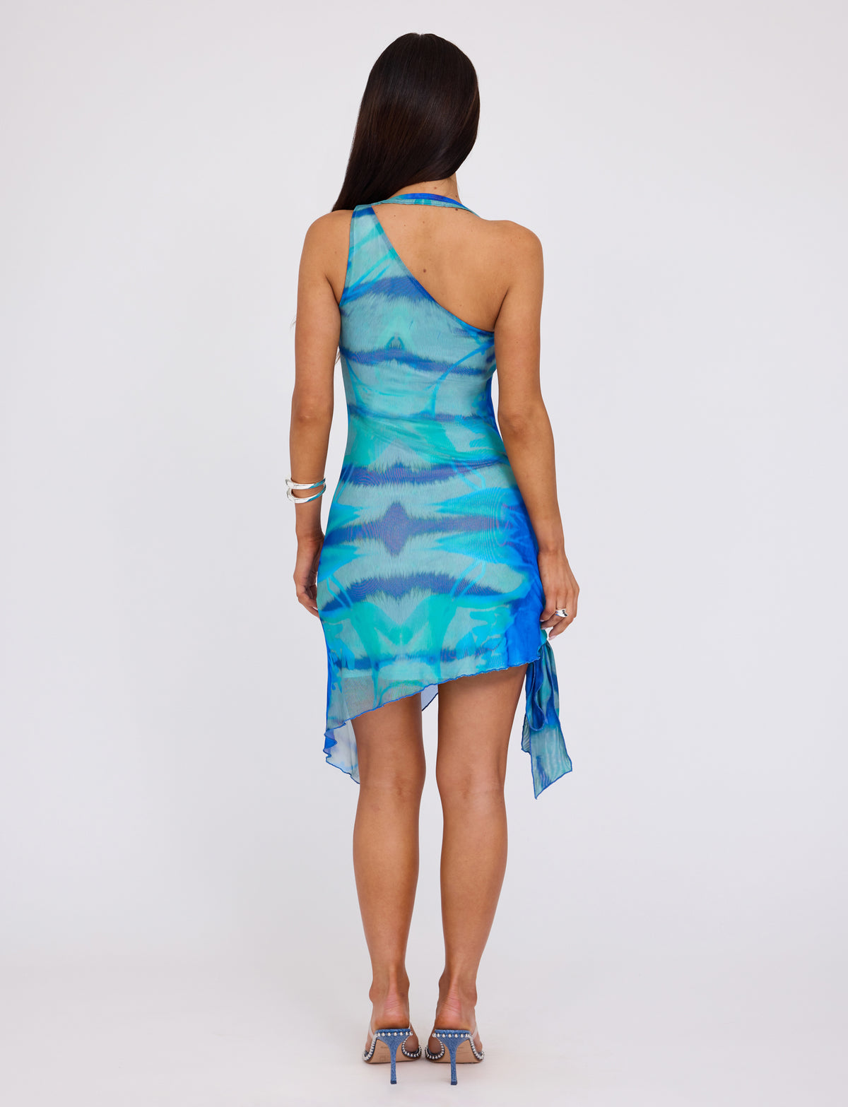 A person with long dark hair, in a sleeveless, asymmetrical TENLEY DRESS - MULTI : ABSTRACT TIE DYE from TIGERMIST with a draped hem, stands with their back to the camera. They also wear blue high-heeled sandals and a silver bracelet on their left wrist.