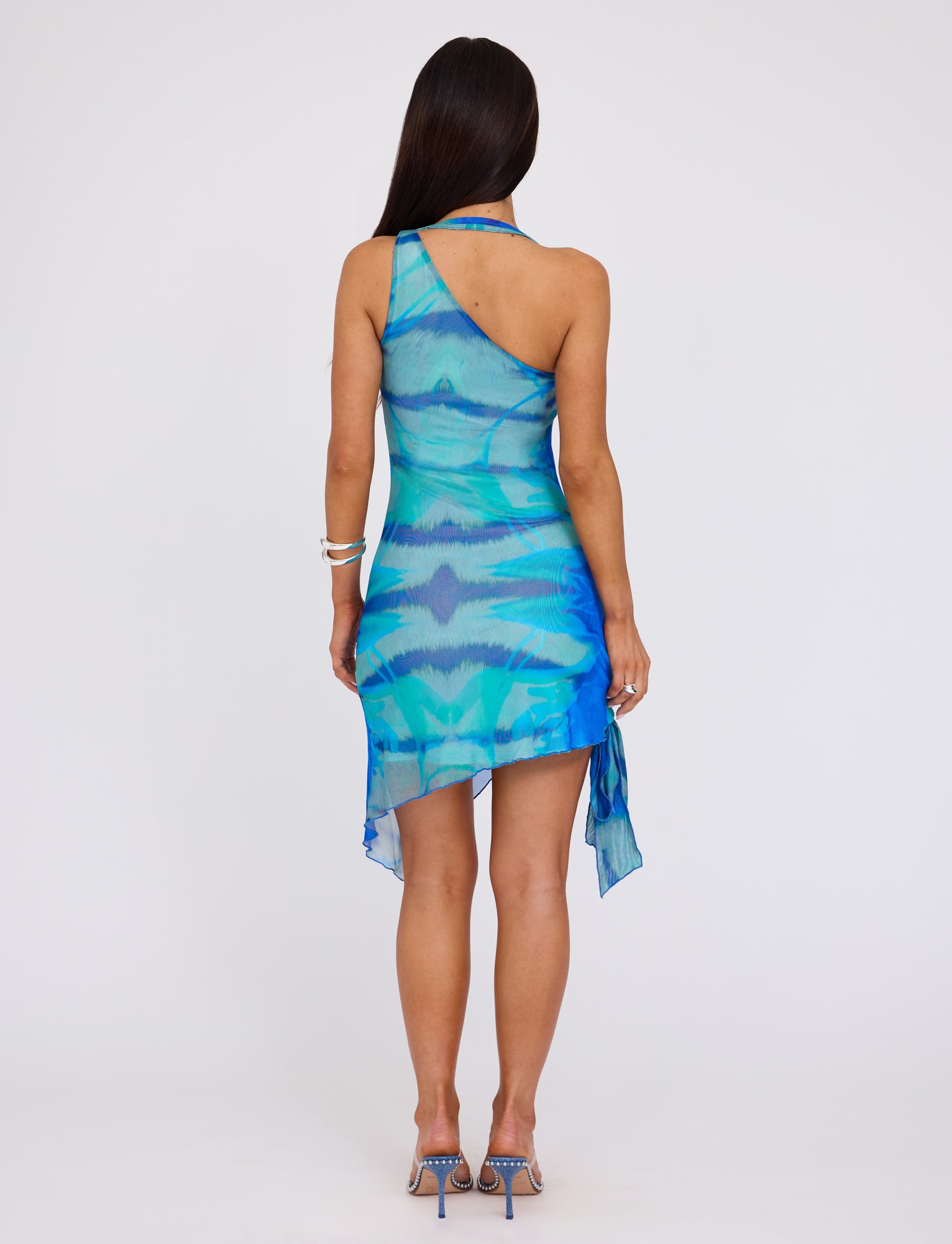 A person with long dark hair, in a sleeveless, asymmetrical TENLEY DRESS - MULTI : ABSTRACT TIE DYE from TIGERMIST with a draped hem, stands with their back to the camera. They also wear blue high-heeled sandals and a silver bracelet on their left wrist.