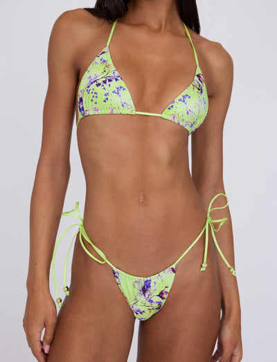 A woman models the Tigermist AVE Bikini Top in a neon green with a purple and black marble pattern. The bikini top features thin straps that tie around the neck and back, and the bottoms have side ties. The background is plain and light-colored.