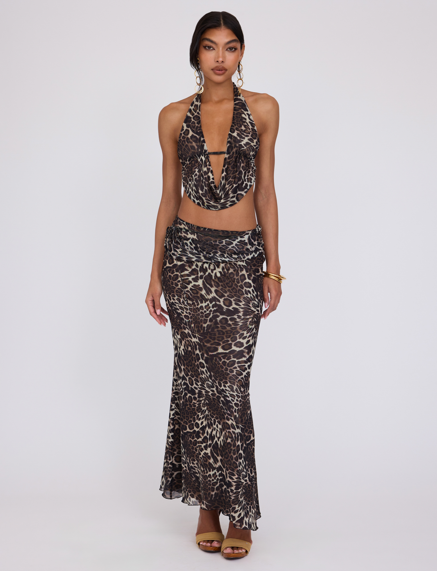 A woman with long dark hair wears the TIGERMIST PEARL TOP - MULTI : LEOPARD, featuring a plunging halter neckline, paired with a matching long skirt, gold earrings, bangles, and brown heels against a plain background. She faces forward neutrally.