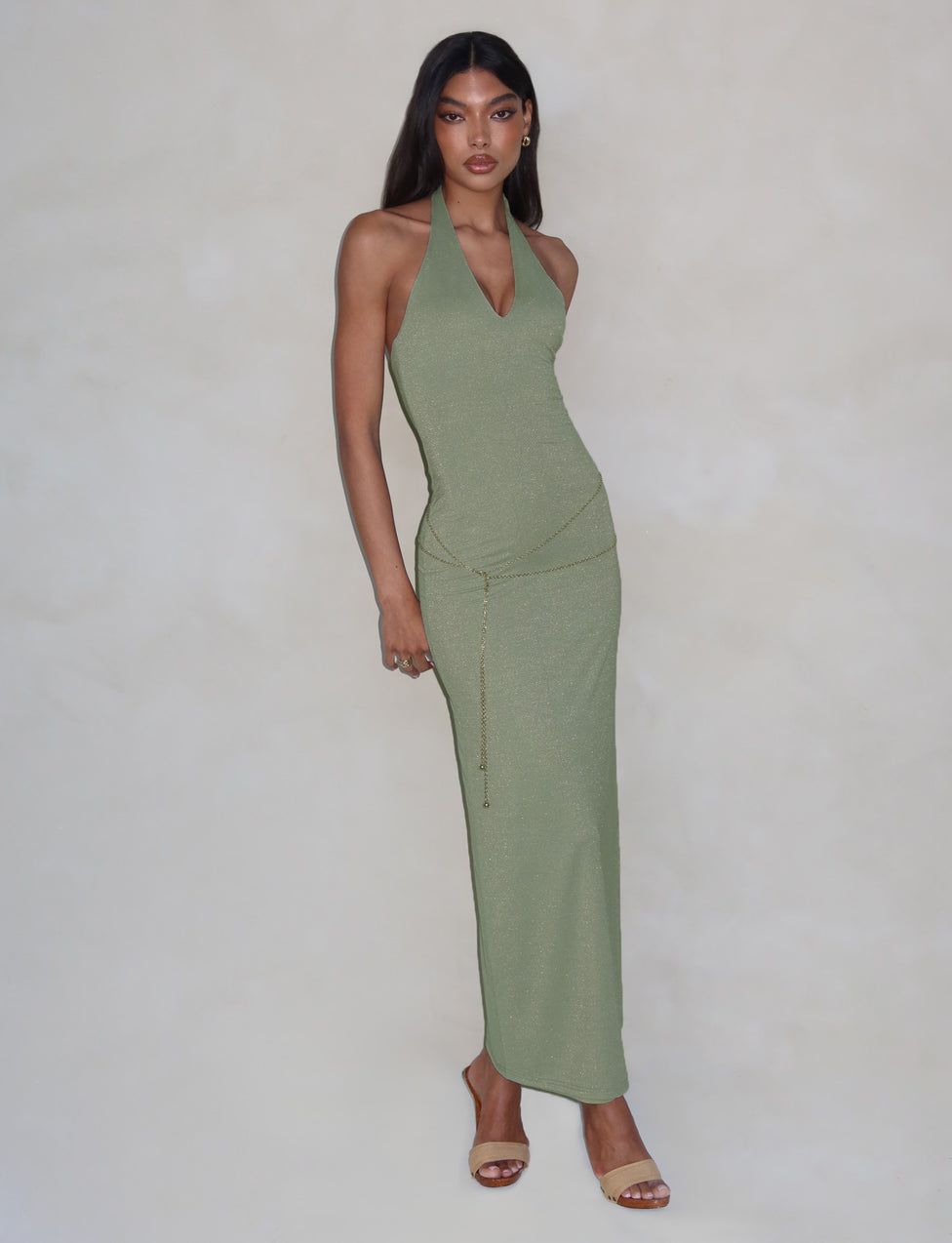 A woman stands against a plain backdrop wearing a long, form-fitting TIGERMIST Opeli Maxi Dress in Green Sparkle, accented with a gold chain belt. She has long, straight hair and is wearing heeled sandals. With a relaxed posture, she gazes directly at the camera.