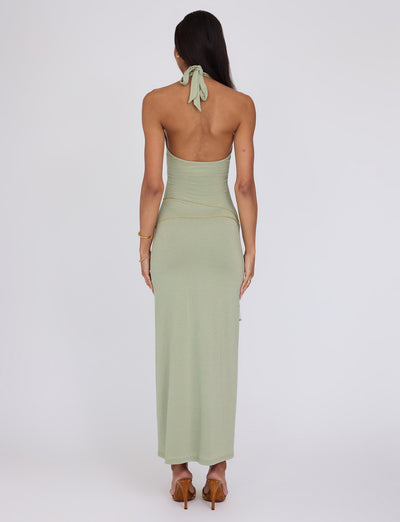A person with long, dark hair is standing with their back to the camera, wearing a light green Opeli Maxi Dress - Green Sparkle from Tigermist that ties behind the neck. They appear to be wearing high-heeled sandals and a gold chain belt around their waist. A bracelet adorns their left wrist against the plain white background.