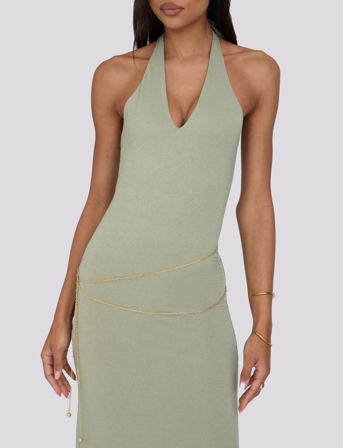 A woman is wearing the sleeveless, halter-neck OPELI MAXI DRESS - GREEN SPARKLE by TIGERMIST, featuring a deep neckline. She accessorizes with a thin gold chain belt draped around her waist and a delicate gold bracelet on her left arm. The background is plain light gray.