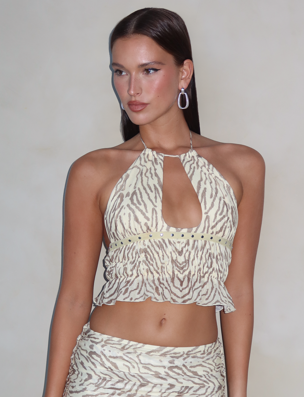A person with long, dark hair poses against a neutral background. They are wearing the TIGERMIST RUMER TOP - YELLOW : ZEBRA, a sleeveless, halter-neck top featuring adjustable bust panels and a keyhole cutout, along with the matching skirt in a yellow zebra print pattern. The person completes their look with large white hoop earrings.