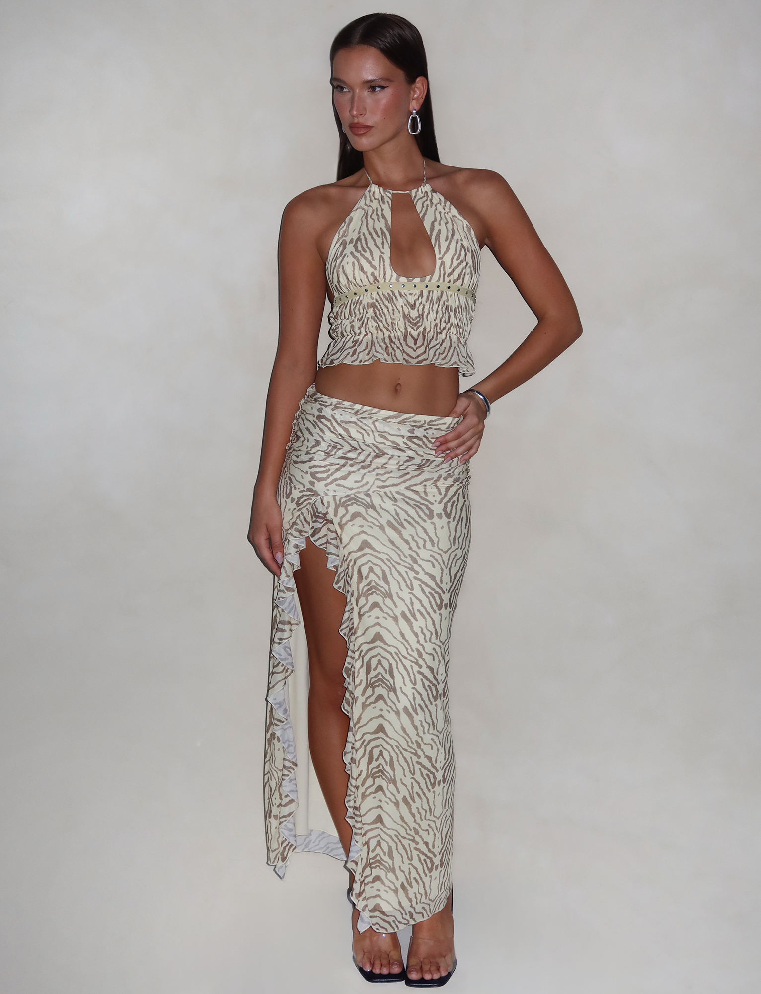 A woman stands against a neutral background, wearing a beige and white zebra-print halter crop top paired with the TIGERMIST RUMER SKIRT in yellow zebra print. She accessorizes with hoop earrings and a bracelet. Her hair is styled straight, and she is wearing heels.