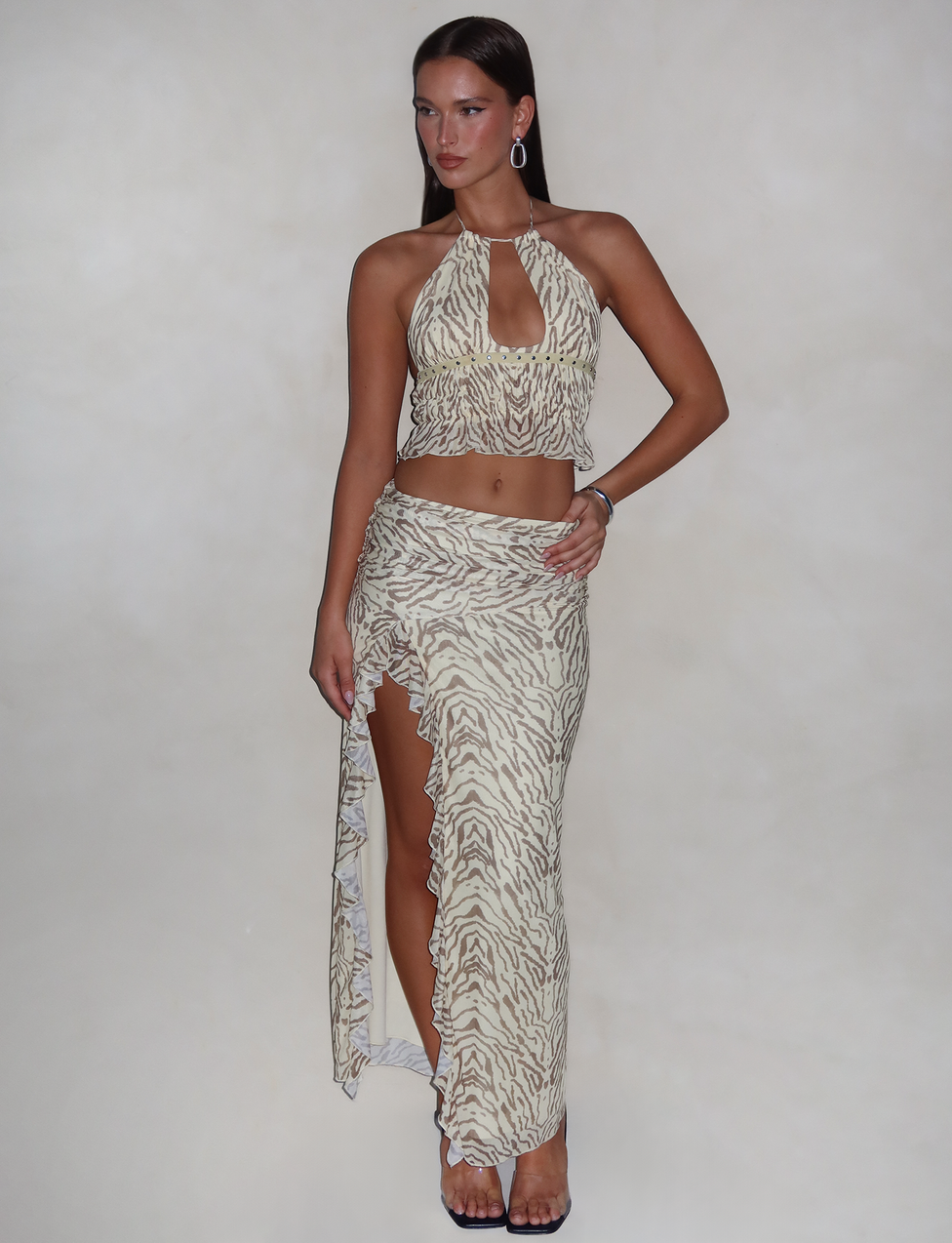 A woman stands against a neutral background, wearing a beige and white zebra-print halter crop top paired with the TIGERMIST RUMER SKIRT in yellow zebra print. She accessorizes with hoop earrings and a bracelet. Her hair is styled straight, and she is wearing heels.