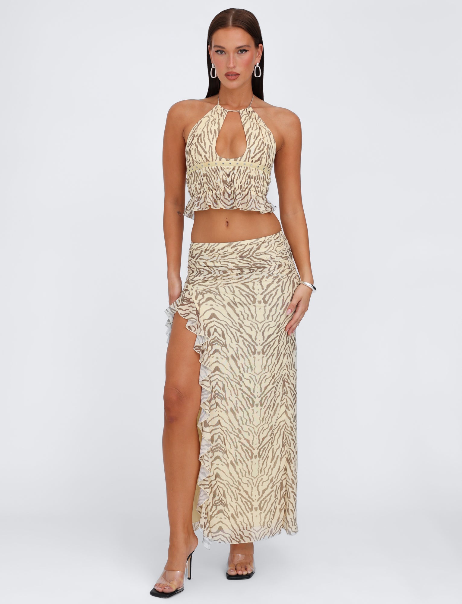 A woman stands confidently in a stylish, sleeveless halter top cropped to reveal her midriff, paired with the TIGERMIST RUMER SKIRT - YELLOW : ZEBRA high-slit maxi skirt. She’s accessorized with hoop earrings, a bracelet, and black high-heeled sandals.