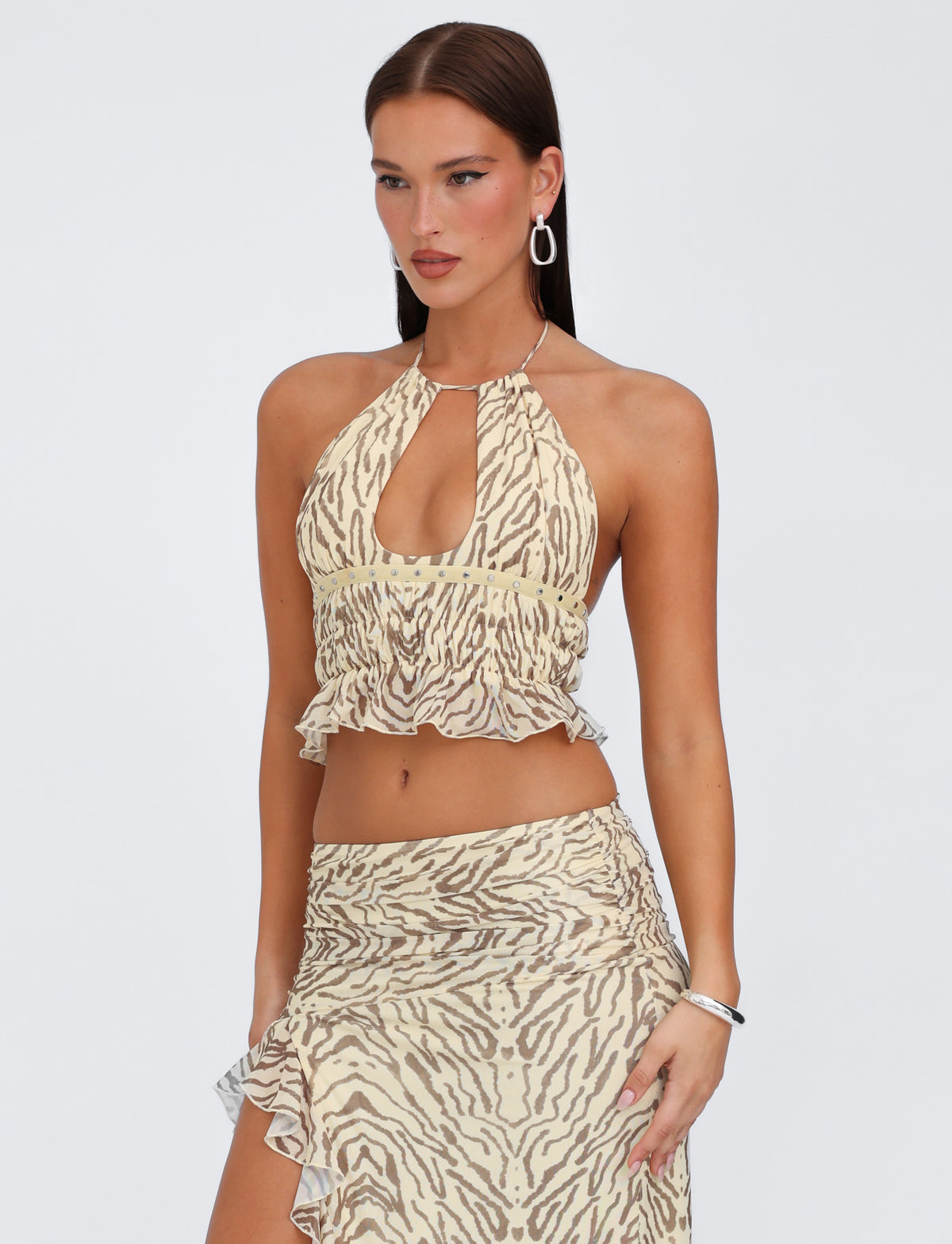 Dressed in the TIGERMIST RUMER TOP - YELLOW : ZEBRA, a woman with long brown hair and hoop earrings models the beige and brown animal print halter top featuring adjustable bust panels and a front keyhole cutout. She pairs it elegantly with a matching high-waisted skirt that boasts a thigh-high slit and ruffled hem as she poses against a plain, light gray background.