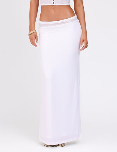 A person models the BENTON SKIRT - WHITE by TIGERMIST, which is a floor-length white maxi skirt featuring a high slit on one side. The outfit is accessorized with a gold bracelet on one wrist. The skirt showcases a fitted design, smooth lightweight fabric, and a delicate lace waistband that adds an elegant touch.