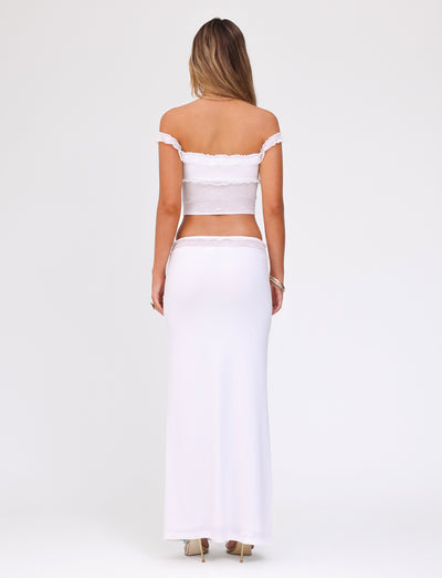 A woman stands with her back to the camera, wearing the TIGERMIST BENTON SKIRT in white, which is a floor-length piece characterized by an off-the-shoulder design and a fitted lace waistband. The skirt flows gracefully down to her heels. She has long hair and is standing against a plain white background.