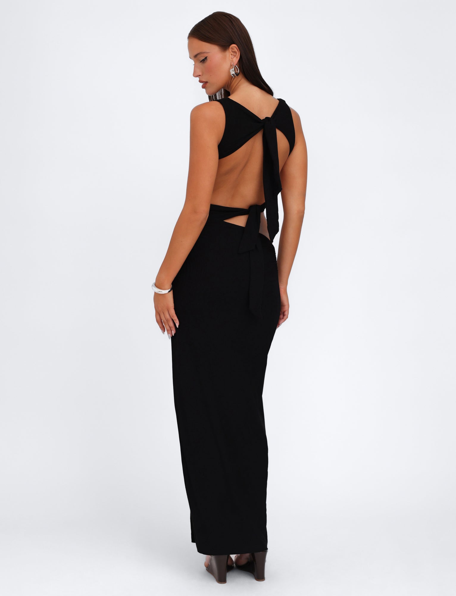 A woman with long brown hair is wearing the TIGERMIST CASPIAN DRESS in black, featuring a bodycon fit, sleeveless design, maxi length skirt, open back, and bow ties at the neck and lower back. She is also accessorized with large earrings and a bracelet, standing sideways while looking over her shoulder.