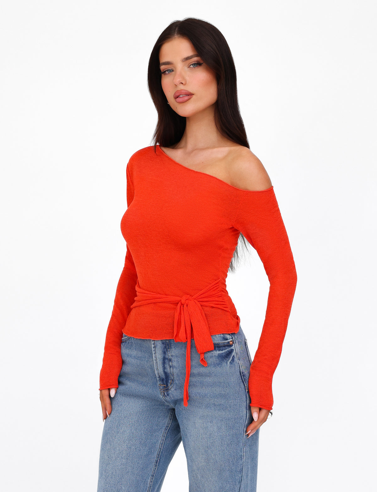 A woman with long, dark hair poses confidently in a fitted silhouette, wearing the TIGERMIST BRYNA JUMPER - RED off-the-shoulder long-sleeve top paired with blue jeans. The background is plain white.