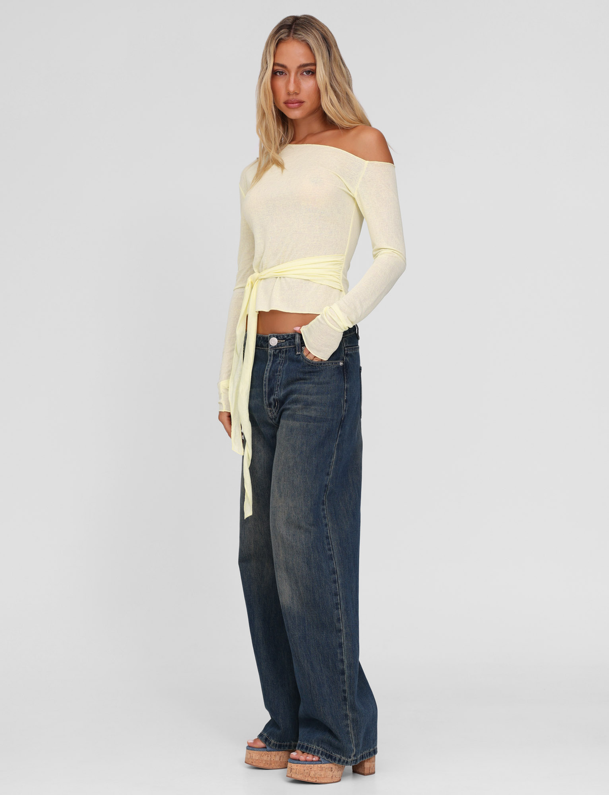 A woman stands against a plain background, wearing the TIGERMIST BRYNA JUMPER in pale lemon and pale yellow, styled with wide-leg dark blue jeans and beige platform sandals.
