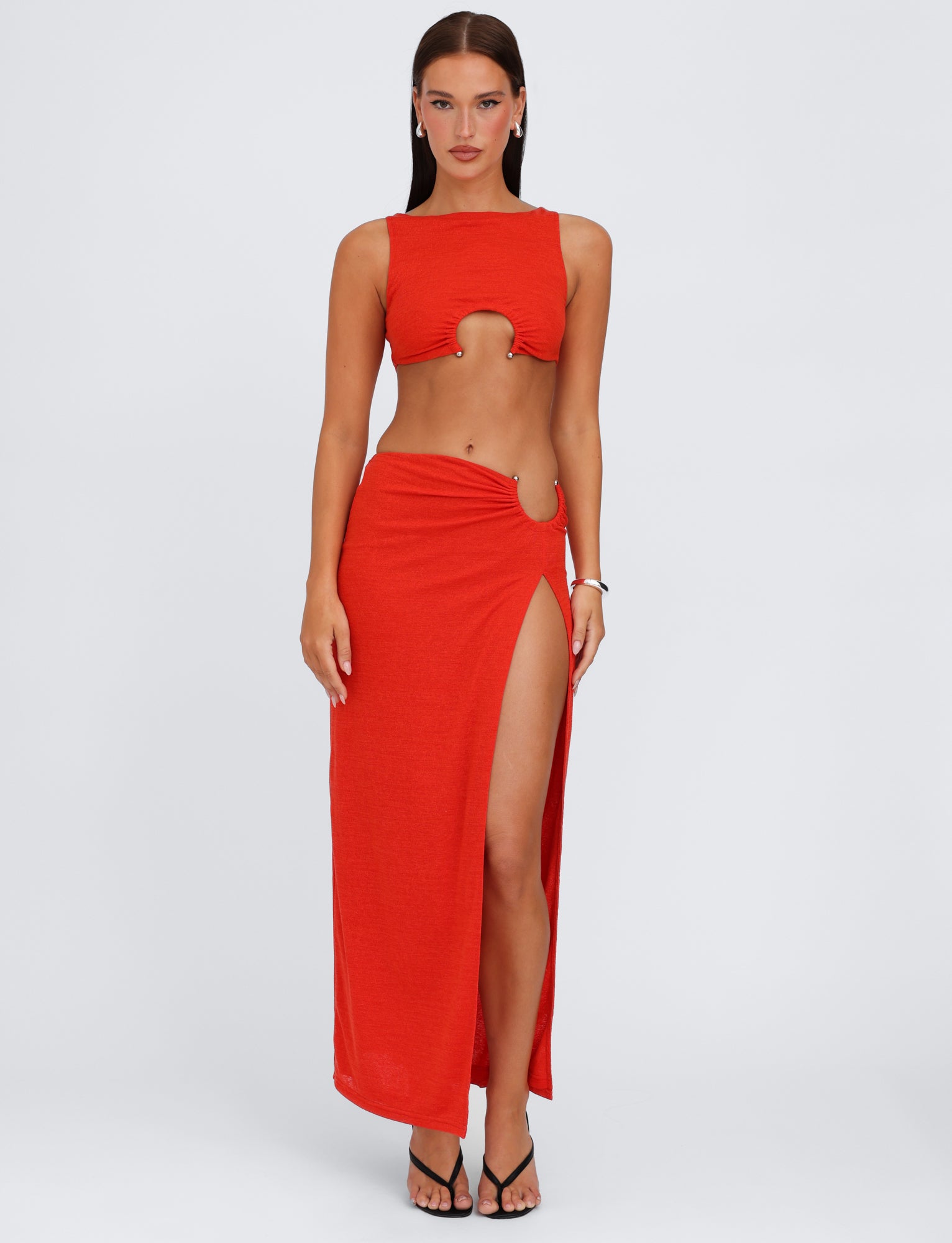 A woman stands against a white background, wearing the vibrant red PAXTON TOP - RED: OMBRE from TIGERMIST. The sleeveless mesh tank top features a circular cutout at the chest and has a cropped length, beautifully complementing the matching high-waisted bodycon skirt that boasts a high slit on one side. She pairs the outfit with black sandals.