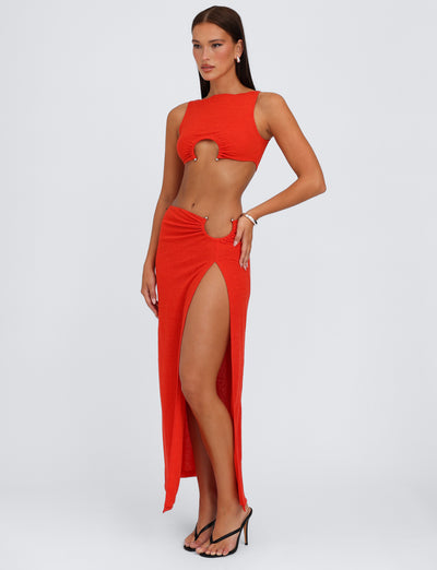A woman poses in the stunning TIGERMIST PAXTON TOP - RED : OMBRE, a striking garment featuring two cut-out sections—one under the bust and another at the hip. The top is paired with a matching high-slit skirt, revealing her leg. She complements her look with black high-heeled sandals and jewelry while standing against a plain background.