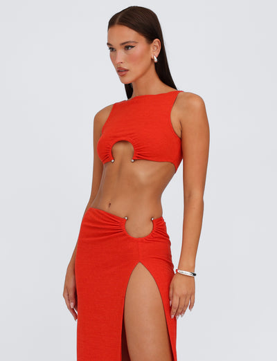 A woman with long brown hair is wearing the PAXTON TOP - RED : OMBRE by TIGERMIST, a sleeveless, bodycon fit orange-red top with cut-out details at the chest and waist, reminiscent of a red mesh tank top. The high slit on her skirt adds flair. She accessorizes with hoop earrings and a bracelet while standing against a plain light background.
