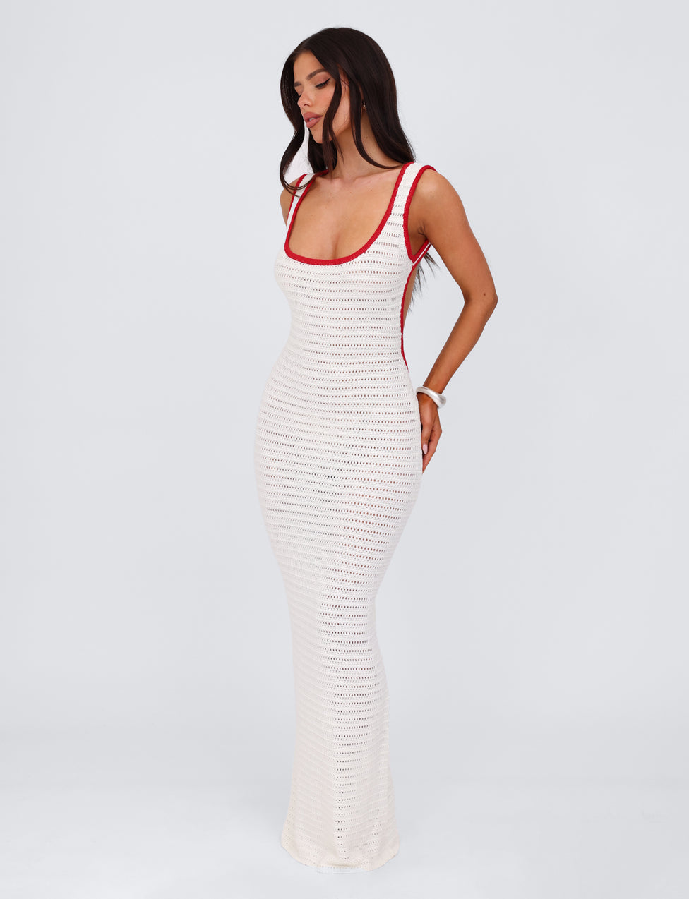 A woman stands against a plain background wearing the MARLEIGH DRESS - WHITE by TIGERMIST. The fitted, sleeveless crochet maxi dress features elegant red trim along the neckline and armholes. Her long, straight hair cascades over her shoulders as she poses with her eyes closed, slightly tilting her head to reveal the sophisticated low back of the dress.