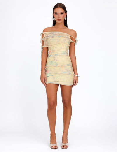 A woman stands against a plain white background, wearing the TIGERMIST KERR DRESS - YELLOW : DELICATE FLORAL, featuring tied straps on the sleeves and ruched detailing. She pairs it with beige high-heeled shoes, minimal jewelry, and has long, straight hair.