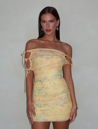 A woman with long, dark hair is wearing the KERR DRESS - YELLOW : DELICATE FLORAL by TIGERMIST. The light yellow, off-shoulder mini-length dress features side cutouts and a pastel floral print. She accessorizes with silver hoop earrings and a bracelet on her right wrist, standing against a plain light gray background.