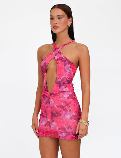 A woman stands in the TIGERMIST KRUZE MINI DRESS - PINK : HIBISCUS, showcasing its high-stretch fabric adorned with bold purple floral patterns. The bodycon fit features a halter neckline and a prominent front cutout. She has long, straight hair and accessorizes with gold hoop earrings and a gold bracelet. Her expression is neutral as she poses against a plain white background.
