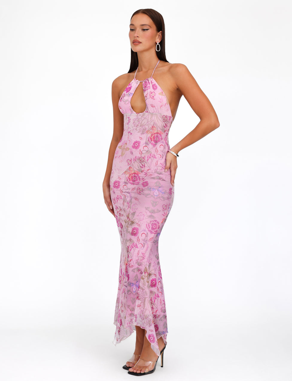 A woman stands against a plain white background, wearing the TIGERMIST COSETTE DRESS - PINK : DELICATE FLORAL, featuring a floor-length, multi-wear halter neckline and front cut-out design. She accessorizes with hoop earrings and a bracelet, completing the look with black high-heeled shoes.
