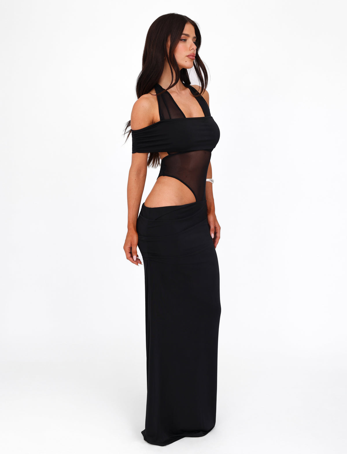 A woman with long dark hair is wearing the elegant TIGERMIST EMBERLAIN DRESS - BLACK, which features a sheer mesh panel bodice with cut-out details on the sides and shoulders, as well as a halter neck design. She stands against a white background, posing sideways with a confident expression.
