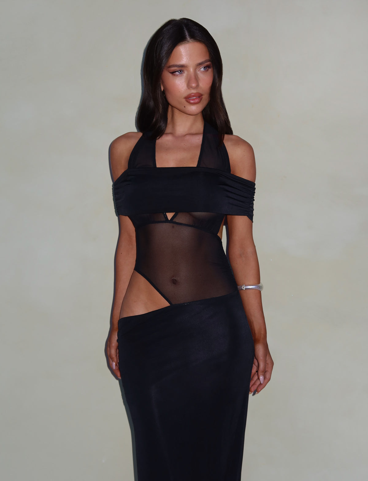 A woman with long, dark hair wears an elegant TIGERMIST EMBERLAIN DRESS in black, featuring a halter neckline and sheer mesh panel midsection, showcasing her arms and shoulders. She poses confidently against a plain, neutral background, accessorizing with a silver bracelet.